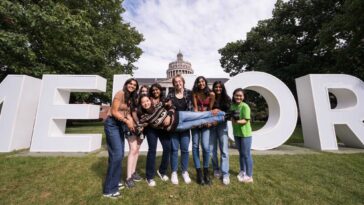 Six University of Rochester students hold up a seventh in front of the giant letters on the quad during Meliora Weekend 2023.