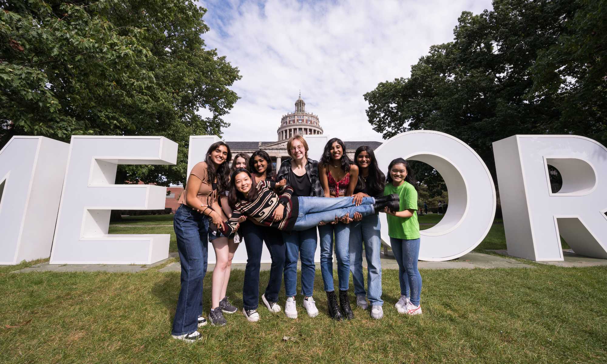 Six University of Rochester students hold up a seventh in front of the giant letters on the quad during Meliora Weekend 2023.