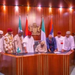 President tinubu signing a document at a desk with nigerian officials standing behind him.
