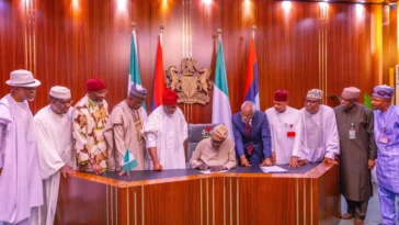 President tinubu signing a document at a desk with nigerian officials standing behind him.