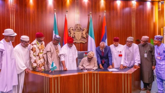 President tinubu signing a document at a desk with nigerian officials standing behind him.
