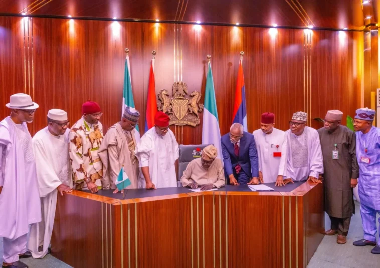 President tinubu signing a document at a desk with nigerian officials standing behind him.