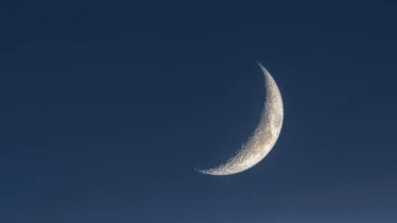 A thin crescent moon glowing in a dark blue twilight sky.