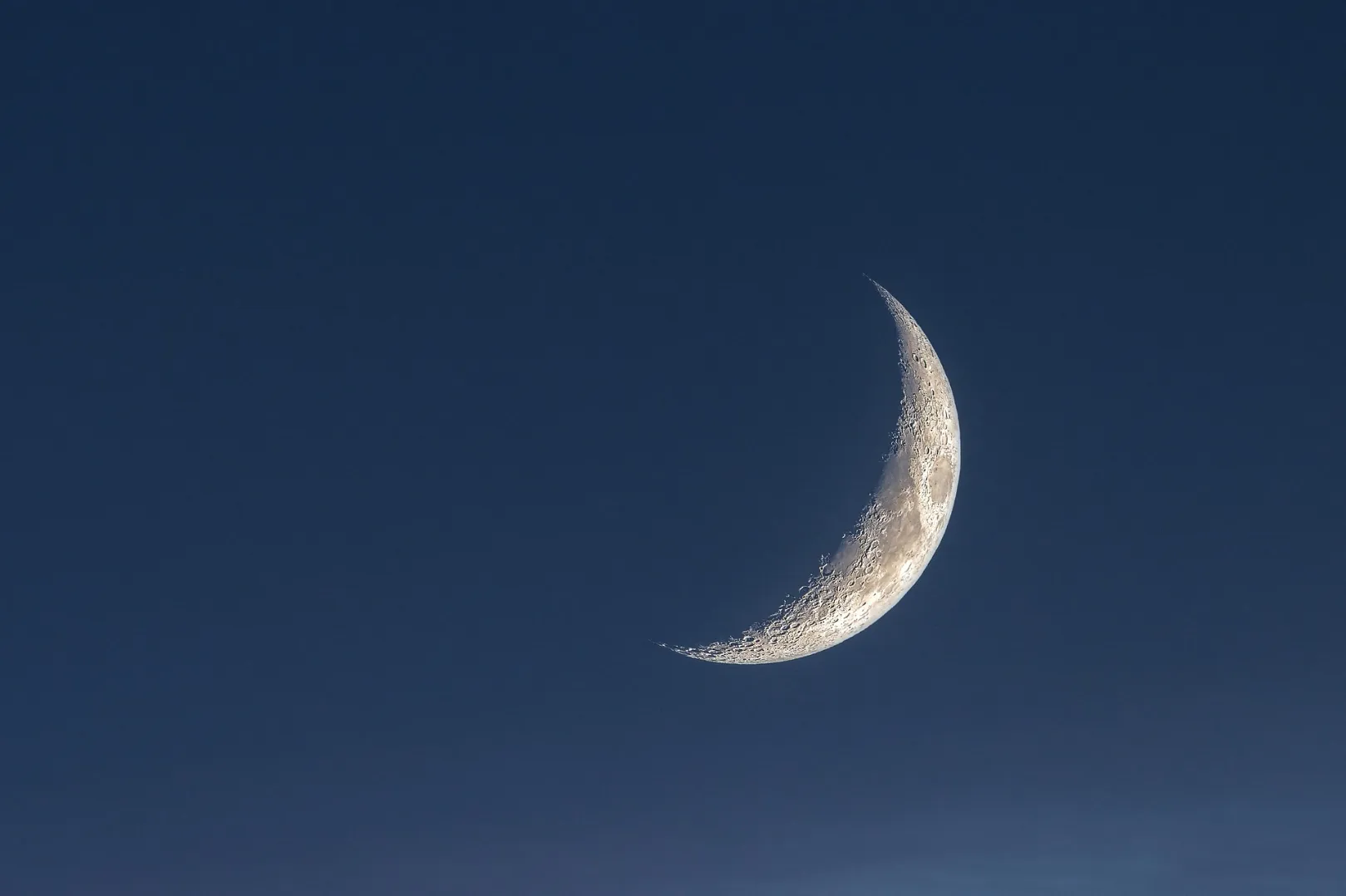 A thin crescent moon glowing in a dark blue twilight sky.